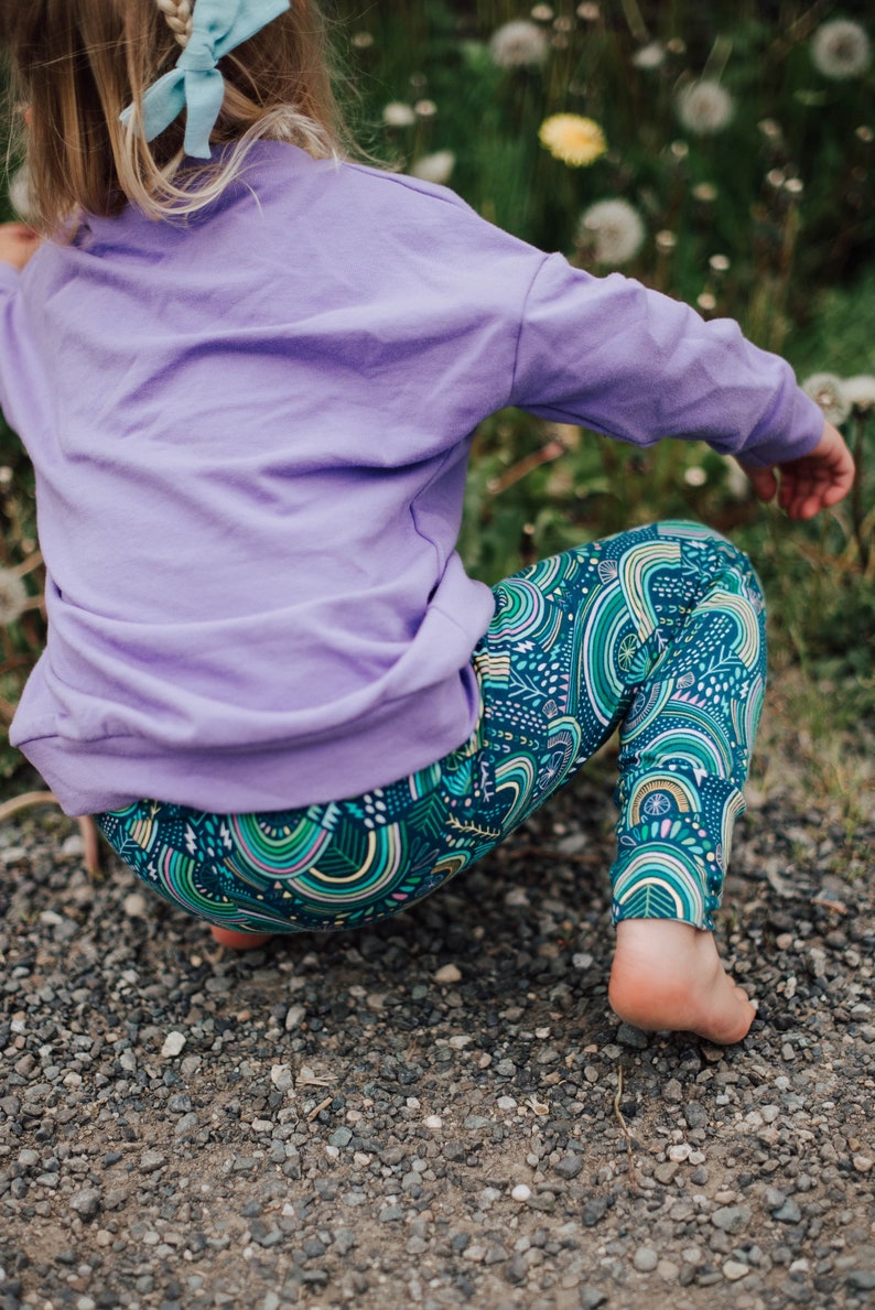 May include: A child in a lavender sweatshirt and patterned leggings. The leggings have a teal background with a repeating design of green, yellow, and white arches and lines. The child is squatting on a gravel surface.