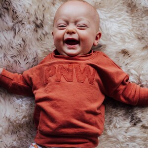 May include: A baby wearing a rust-colored sweatshirt with "PNW" embroidered on the front, and patterned pants with sun graphics, smiles broadly while lying on a fluffy, cream-colored rug.