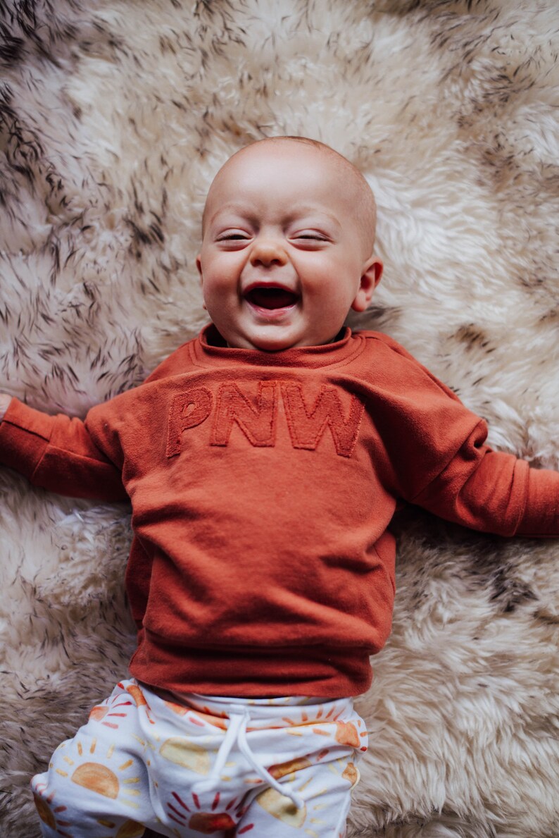 May include: A baby wearing a rust-colored sweatshirt with "PNW" embroidered on the front, and patterned pants with sun graphics, smiles broadly while lying on a fluffy, cream-colored rug.