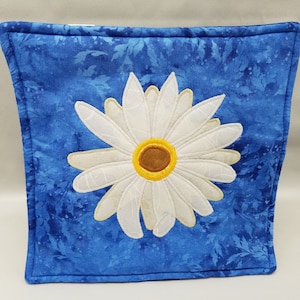 May include: A blue pillow with a white daisy embroidered on it. The daisy has a yellow center and is surrounded by a blue and white floral pattern.