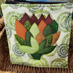 May include: A square, quilted fabric item featuring an artichoke design. The artichoke is composed of various shades of green, orange, and brown, with a detailed, layered appearance. The background fabric has a white base with a green, circular pattern.