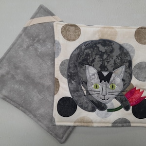 May include: A square quilted potholder featuring a gray cat appliqué holding a pink flower on a cream background with large brown, gray, and black polka dots. The reverse side is a solid mottled gray fabric with a small beige hanging loop.