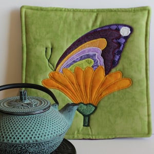 May include: A teal cast iron teapot with a textured hobnail pattern and a curved handle sits in front of a square green fabric wall hanging featuring an appliqued orange flower and a purple, blue, and white butterfly with stitched details.