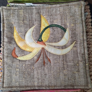 May include: A square fabric coaster featuring a textured brown background with a central appliqué of a stylized flower. The flower has yellow and cream-colored petals, a dark green curved stem, and three thin brown stamens extending downward.
