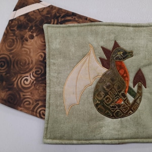 May include: A square quilted potholder featuring a green fabric background with an appliqued dragon design. The dragon has cream-colored wings, a dark green body with a patterned texture, and an orange chest. A second brown fabric piece is visible.