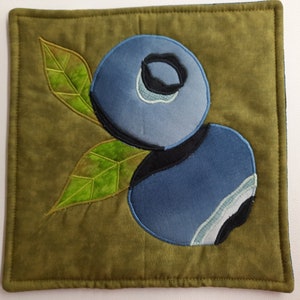 May include: A green fabric coaster with two embroidered blueberries. The blueberries are blue and have a black outline. The leaves are green and have a light green outline.