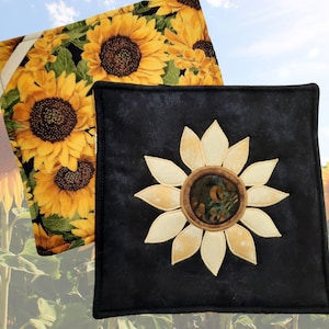 May include: Two square fabric potholders. One features a vibrant sunflower print on a black background. The other is solid black with a central fabric appliqué of a sunflower, showcasing cream-colored petals and a dark, textured circular center.