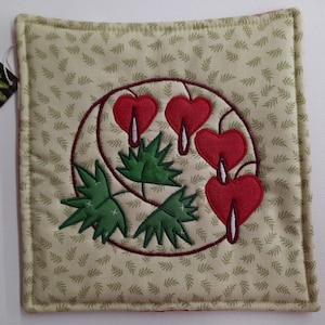 May include: A green and red quilted hot pad with a design of bleeding hearts. The background is a light green with a small leaf pattern.