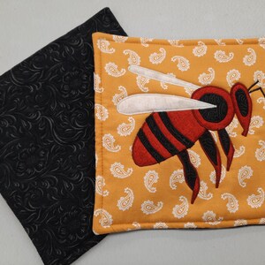 May include: Two square pot holders. One is black with a floral pattern. The other is mustard yellow with a white paisley pattern and an embroidered bee with red, black, and white details.