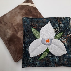 May include: Two quilted pot holders, one brown with white trim and one blue with a white flower design. The flower is a white trillium with a green stem and orange center.