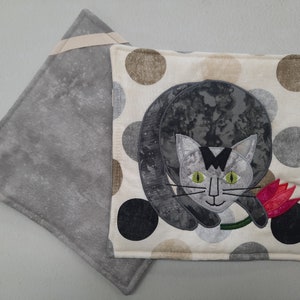May include: A gray and white pot holder with a gray cat holding a pink flower on a white background with brown and black polka dots.