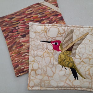 May include: Two square fabric pot holders. One has a hummingbird applique with a red throat and a black beak. The other has a brown and red abstract pattern. Both have a cream-colored border.