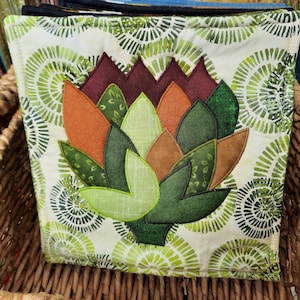 May include: A green, orange, and brown quilted pot holder with a flower design on a white background with green circles.
