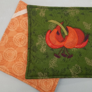May include: Two square quilted pot holders. One is orange with a circular pattern. The other is green with an embroidered orange flower design. The green pot holder has a floral pattern in the background.