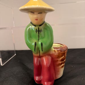 May include: A ceramic figurine of a person wearing a yellow conical hat, a green jacket with black line details, and red trousers. The figure is leaning against a brown, textured cylindrical basket-like container with a cream-colored interior.