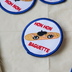 May include: Four embroidered iron-on patches featuring a cartoon baguette wearing a beret and the text "Hon Hon Baguette".