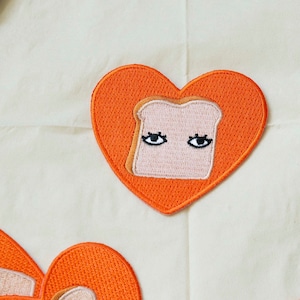May include: Orange heart-shaped patches with embroidered images of toast with eyes. The toast is light brown and the eyes are black.