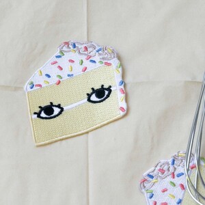 Puede incluir: Tres parches bordados para planchar de forma de rebanadas de pastel amarillo con glaseado blanco y chispas de colores. Cada rebanada tiene dos ojos negros con pupilas blancas.