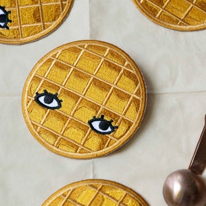 Puede incluir: Cuatro parches bordados en forma de gofre amarillo con ojos y pestañas negras. Los parches están sobre una superficie blanca con un batidor de alambre y una cuchara de metal.