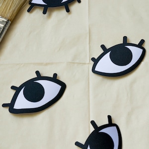 May include: Four black paper eye cutouts with white pupils and eyelashes on a beige surface. A wooden paintbrush and a black silicone spatula are also on the surface.