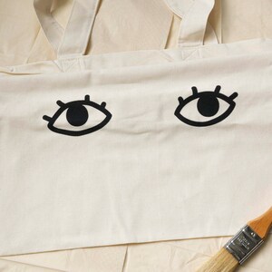 Puede incluir: Una bolsa de lona blanca con dos ojos negros impresos en la parte delantera. Los ojos tienen pestañas y pupilas negras. La bolsa tiene dos correas blancas.