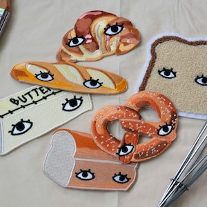 May include: Six embroidered patches featuring various baked goods with eyes. The patches include a croissant, a loaf of bread, a pretzel, a baguette, a stick of butter, and a loaf of sourdough bread. The patches are all on a beige fabric background.