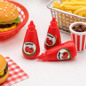 May include: Red ketchup bottles with white labels featuring tomato illustrations. The bottles are positioned near a miniature burger in a red basket, fries in a metal basket, and a drink in a red and white striped cup. The scene evokes a fast-food theme.