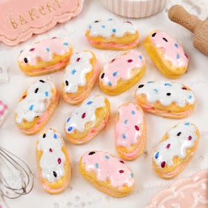 May include: A collection of miniature faux eclairs featuring golden-brown pastry shells filled with pink or white cream, topped with glossy white or pink icing and colorful sprinkles. These small decorative food replicas are arranged on a surface.