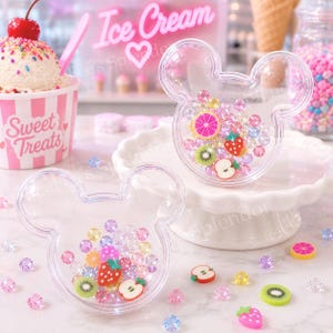 May include: Two clear, Mickey Mouse-shaped containers filled with colorful gems and fruit-shaped charms. The containers sit on a white surface, surrounded by loose gems and fruit slices. A pink ice cream cup with the words "Sweet Treats" is visible.