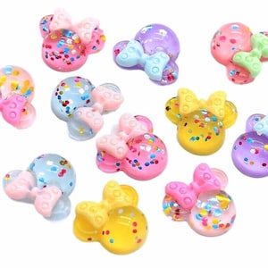 May include: A collection of small, translucent resin cabochons shaped like mouse heads with bows. Each piece features colorful embedded glitter in various shades including pink, blue, yellow, and purple, with matching solid-colored bows.