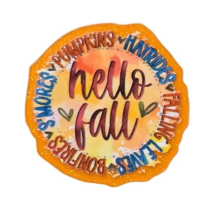 May include: A glittery orange sticker with the words "hello fall" in cursive, surrounded by fall-themed words like "pumpkins" and "bonfires." The background has watercolor-like orange and yellow hues.