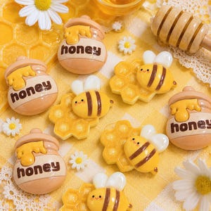 May include: A collection of small, flat-backed resin charms featuring honey pots with the word honey printed on them, and yellow bees resting on honeycomb shapes. The charms are interspersed with small white daisy flowers on a yellow checkered fabric.