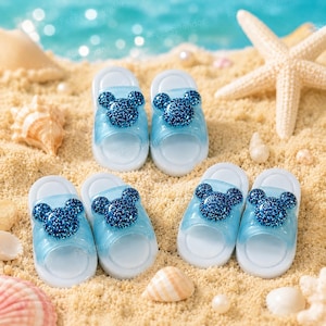 May include: Six pairs of light blue children's sandals with a glittery dark blue Mickey Mouse design on the top. The sandals are arranged on a sandy surface with seashells and a starfish.