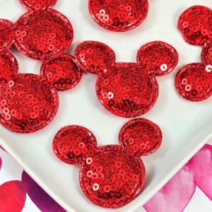 May include: A collection of red sequin patches shaped like mouse heads with two circular ears and a round face. Each patch is covered in small, shiny red sequins that reflect light, creating a textured and sparkling appearance on the surface.