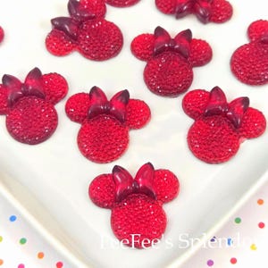 May include: Red Minnie Mouse-shaped embellishments with sparkling details and a glossy bow. These decorative pieces are arranged on a white surface. The image includes the text "FeeFee's Splendor".