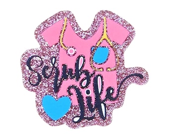 May include: A pink scrub top shaped sticker with a glittery border. It features a yellow stethoscope, a blue heart, and the black cursive text Scrub Life. The design is centered on a pink background with a textured, sparkling glitter finish.