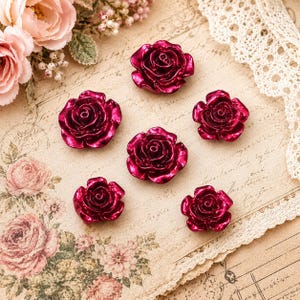May include: Six individual rose-shaped embellishments in a deep, metallic magenta finish. Each piece features layered, sculpted petals arranged in a circular bloom pattern, showcasing a glossy, reflective surface texture throughout the set.