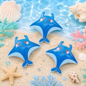 May include: Three blue manta ray figurines with light blue undersides and smiling faces are arranged on a sandy surface with seashells, starfish, and coral. The figurines have a cartoonish design.