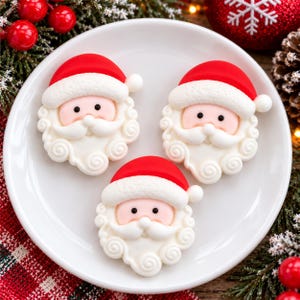 May include: Three identical Santa Claus face decorations arranged on a white plate. Each figure features a bright red hat with a textured white brim, a pale pink face with small black eyes, and a detailed white beard with swirling patterns.