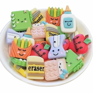 May include: A white bowl filled with assorted school-themed flatback charms featuring smiling faces. The collection includes green notebooks, orange crayon boxes, white glue bottles, red apples with rulers, purple pencil sharpeners, and pink calculators.