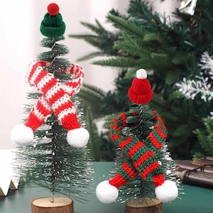 May include: Two miniature Christmas trees decorated with knitted hats and scarves. One tree has a green hat and a red and white striped scarf with white pom-poms. The other tree has a red hat and a green and red striped scarf.