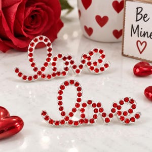 May include: Two silver-toned metal script word pins spelling the word Love, each encrusted with small, round, vibrant red rhinestones. The cursive lettering features a continuous, flowing design with a polished metallic finish and bright red stones.