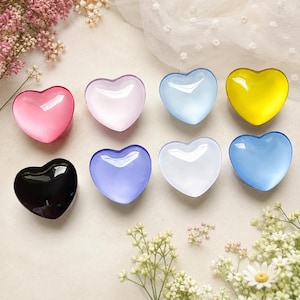 May include: A collection of eight smooth, heart-shaped objects in various colors including pink, light pink, light blue, yellow, black, purple, white, and medium blue. Each heart has a glossy, reflective surface and a rounded, puffed shape.