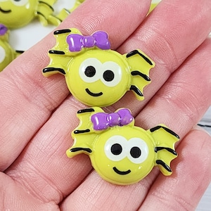 May include: Two yellow bat-shaped embellishments with black details and purple bows. Each has a smiling face with black eyes and a curved mouth. The embellishments are likely for crafts or decorations.