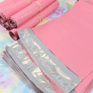 May include: Pink poly mailer shipping envelopes, both rolled and flat, are displayed. The rolled envelopes are secured with rubber bands. The envelopes have a self-sealing adhesive strip. The background is a colorful, tie-dye pattern.