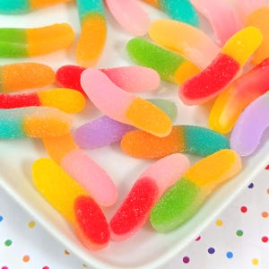May include: A close-up shot of a white plate filled with colorful gummy worms. The gummy worms are in various colors, including red, yellow, green, orange, pink, and purple. The candies have a sugared coating.
