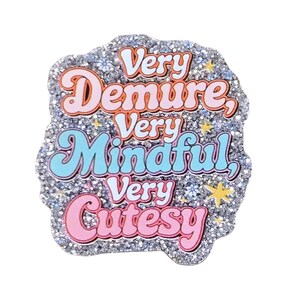 May include: A silver glitter enamel pin with the text Very Demure, Very Mindful, Very Cutesy in orange, blue, and pink lettering. The design includes small yellow star accents and tiny flower details scattered across the sparkling surface.