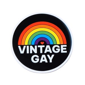 May include: A round sticker with a black background and a rainbow arch design. The text "VINTAGE GAY" is in white, with a small purple heart at the center of the rainbow. The sticker has a white border.
