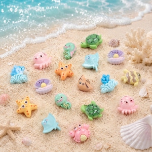 May include: A collection of colorful miniature sea creatures, including starfish, turtles, octopuses, and fish, are arranged on a sandy beach. The figurines are in various shades of pink, green, orange, and blue, creating a playful ocean theme.