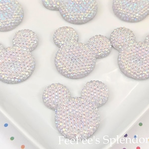 May include: Close-up of several white, iridescent Mickey Mouse-shaped embellishments. Each is covered in small, sparkling crystals. The embellishments are arranged on a white surface. The text "FeeFee's Splendor" is visible.